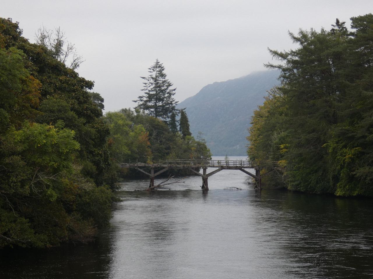 The old wooden bridge at Invermoriston. The Moriston River flows beneath the structure as it makes its way toward the vastness of Loch Ness, visible in the misty background.